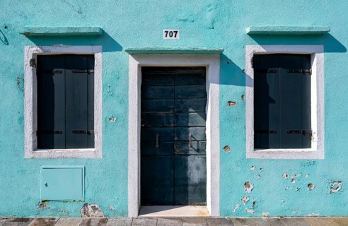 Light blue house facade with entrance door and windows with closed shutters, colourful houses on