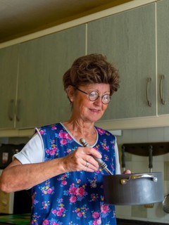 Granny with old smock apron, glasses and wig in the kitchen, holding cooking pot and ladle,