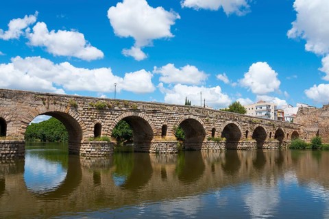 Ancient stone bridge with reflections in the river water, under blue sky with some clouds, Roman
