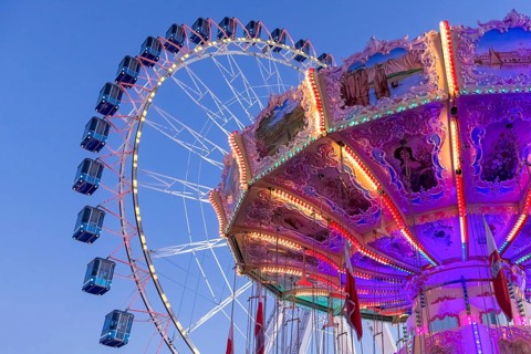 A brightly lit carousel in front of a large Ferris wheel at dusk at a funfair, Wellenflug,