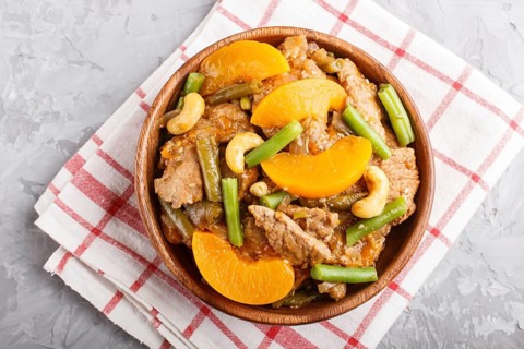 Fried pork with peaches, cashew and green beans in a wooden bowl on a gray concrete background. Top