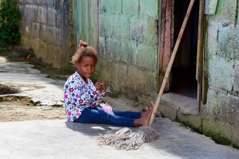 A young girl plays on the ground next to a broom in front of a house in the countryside, between