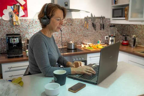 Woman, mid-50s, works from home, with laptop and communicates with colleagues via headset, home