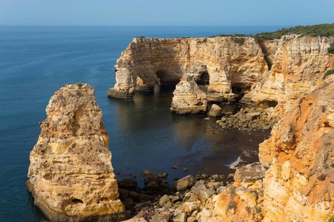 Large, eroded cliffs and rocks stretch along a coastline with calm seas and clear skies, Praia da