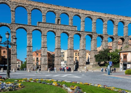 A historic aqueduct with stone arches spans a town with a flowerbed in the foreground under a blue