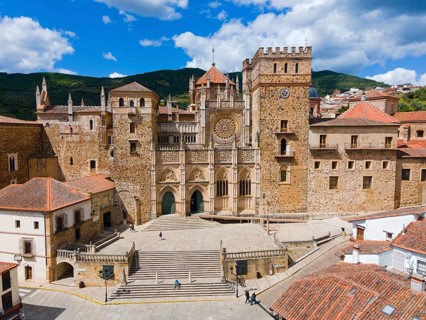 Aerial view of the facade of a monastery with red roofs and wide stairs under a blue sky in Spain.