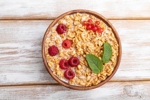 Wheat flakes porridge with milk, raspberry and currant in wooden bowl on white wooden background.