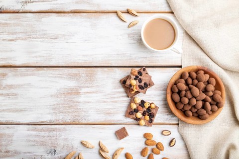 Almond in chocolate dragees in wooden plate and a cup of coffee on white wooden background and