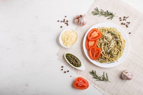 Spaghetti pasta with pesto sauce, tomatoes and cheese on a linen tablecloth on white wooden