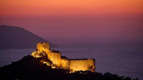 View of an illuminated castle ruin at dusk, surrounded by sea and mountains under a colourful sky,