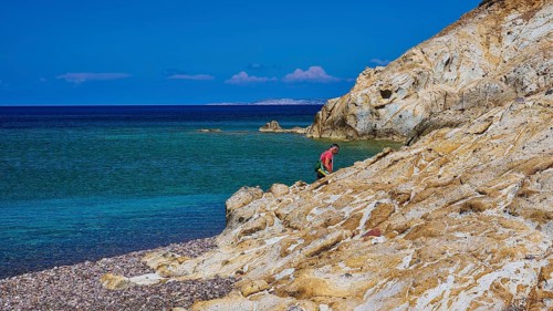 A man climbing on rocks on a coast with clear sky and clear water, Lambi beach, pebble beach,
