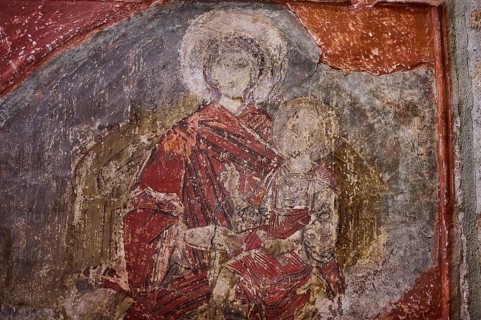 An old fresco shows a holy figure in red robes holding a child, with a weathered background, Church