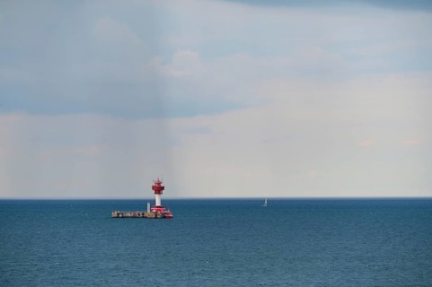 A red lighthouse stands on a calm, blue sea under a slightly cloudy sky, with a small sailing boat