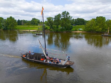 A boat sails with passengers on a calm river, surrounded by green landscape and trees under a