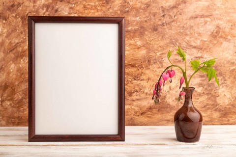 Wooden frame with pink dicentra, broken heart in ceramic vase on brown concrete background. side
