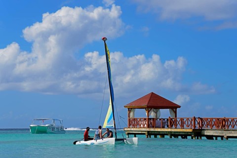 A colourful catamaran sails next to a jetty near a pavilion on the clear sea under a blue sky,