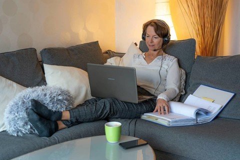 Woman, mid-50s, works from home, with laptop and communicates with colleagues via headset, home