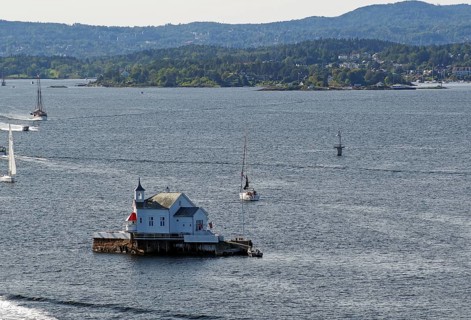 Small lighthouse on an island in the water, surrounded by boats and hills in the distance, Dyna
