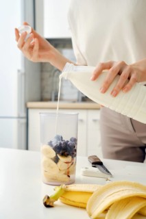 Unrecognizable woman pours milk from bottle into bowl of blender to make a smoothie with black