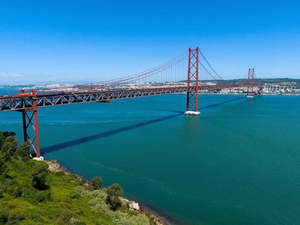 A large red bridge stretches across a wide expanse of water under a clear blue sky, surrounded by