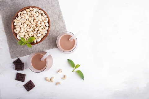 Organic non dairy cashew chocolate milk in glass and wooden plate with cashew nuts on a gray