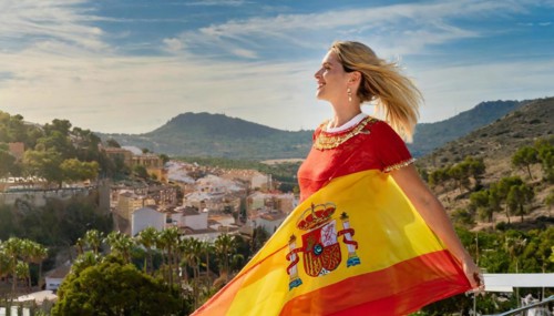 A happy blonde woman holding the flag of Spain, historical town in the background, AI generated, AI