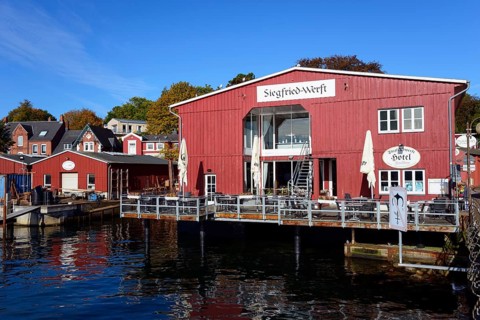 A red wooden building with 'Hotel' lettering on the waterfront under a clear blue sky on a sunny