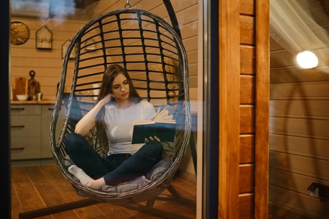 A young woman relaxes in the evening in a wooden house sitting in a hanging chair with a book