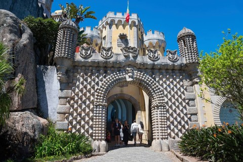 Entrance gate of a castle with decorative stone patterns and plants, visitors are visible, Palácio