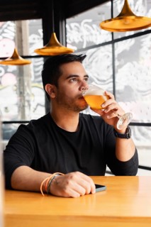 A young man in a black shirt sits at a wooden bar table, enjoying a glass of beer in a stylish