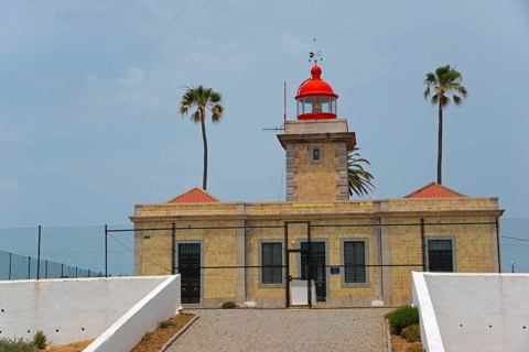 A traditional lighthouse with red roofs and palm trees, surrounded by a coastal landscape, Ponta da