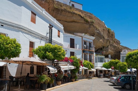 Street with cars and restaurants under rocks with white houses, sunny sky in Setenil de las