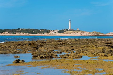 A lighthouse stands on a rocky coast with a blue sky and calm sea in the background, Faro de