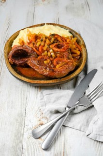 A wooden plate of osso buco served with mashed potatoes, cooked in a tomato and onion sauce with