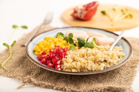 Mixed quinoa porridge, sweet corn, pomegranate seeds and small sausages on white wooden background.