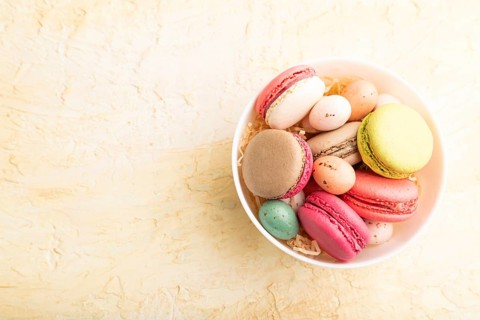 Multicolored macaroons and chocolate eggs in ceramic bowl on beige concrete background. top view,