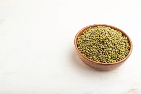 Wooden bowl with raw green mung bean on a white wooden background. Side view, close up, copy space