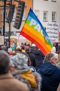 People at a public gathering with a big peace flag 'Pace' and speakers on a stage, Against Right