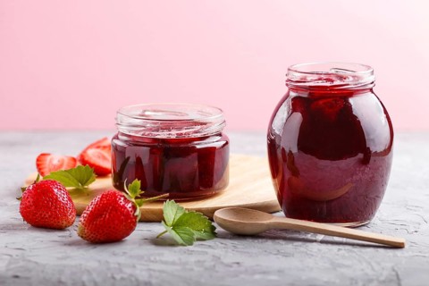 Strawberry jam in a glass jar with berries and leaves on gray concrete background. Homemade, close