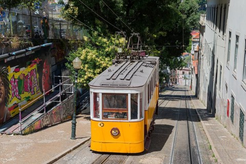 Yellow tram descends past wall art and green trees, funicular railway, Ascensor da Glória, Old