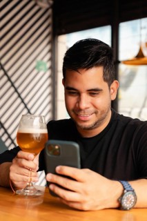 A smiling man in a black shirt enjoys a glass of beer while using his smartphone in a modern bar.