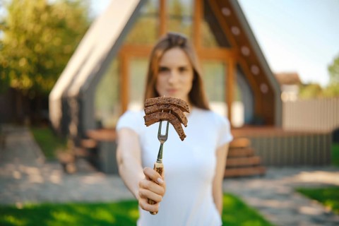 Defocused young woman stretching forward a barbecue fork with slices of steak in her hand