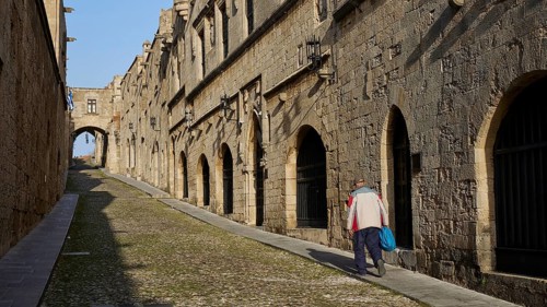 A historic alley with stone buildings, a pedestrian walks along the alleyway in the sunshine,