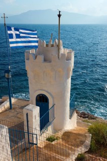 White tower on the coast, with Greek flag in the wind, at blue sea level, Church of Agios Nikolaos