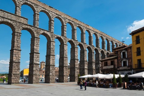 Historic stone aqueduct in a town square with buildings, tables and people, Aqueduct, Segovia,