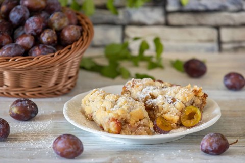 Plum cake on a plate next to a basket with fresh plums in the background, on a white rustic table