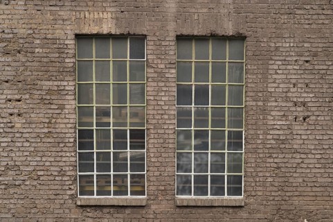 Two large industrial windows with small panes and metal frames are set in an old brick wall,