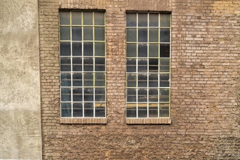 Two large industrial windows with small panes and metal bars are set in an old brick wall, showing