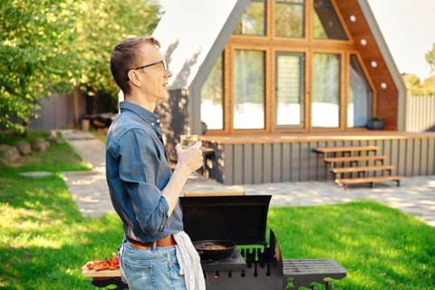 Middle-aged man holds wine glass while standing in profile at the grill in the backyard of a modern