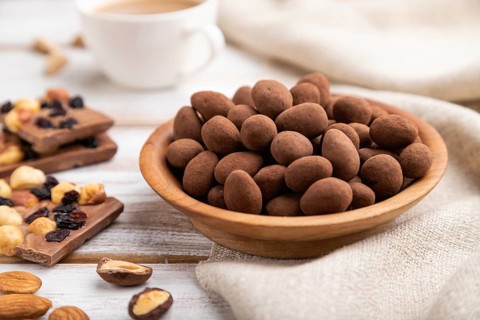 Almond in chocolate dragees in wooden plate and a cup of coffee on white wooden background and